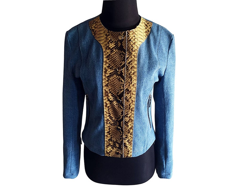 Op de afbeelding: Een blauw denim jack met een slangenhuidpatroon op de voorkant. Het jack heeft een ritssluiting en een opstaande kraag.