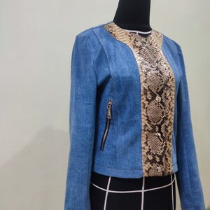 Op de afbeelding: Een blauw denim jack met een slangenvellen paneel aan de voorkant. Het jack heeft een ritssluiting en lange mouwen.