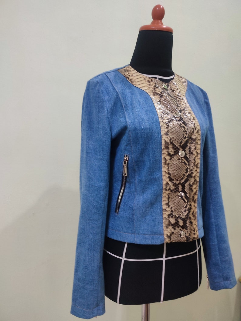 Op de afbeelding: Een blauw denim jack met een slangenvellen paneel aan de voorkant. Het jack heeft een ritssluiting en lange mouwen.