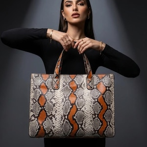 May include: A rectangular tote bag with a snakeskin pattern in shades of gray, orange, and cream. The bag has two top handles and a structured design. The handles and trim are a solid orange color.