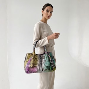 May include: A colorful snakeskin-patterned tote bag with a tassel. The bag features a mix of purple, green, yellow, and gray hues. The handles are black and the bag is held by a person wearing a cream-colored outfit.