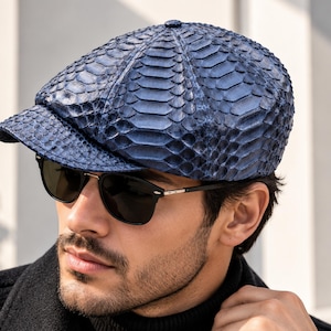 May include: A navy blue newsboy cap featuring a textured, scale-like pattern across the entire surface. The cap has a rounded, multi-paneled crown and a short, stiff brim. The material has a glossy finish that highlights the intricate scale texture.