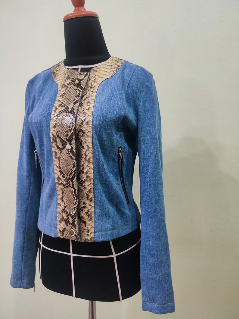 Op de afbeelding: Een blauwe denimjas met een slangenhuidpatroon op de voorkant. De jas heeft een ritssluiting en twee zakken aan de voorkant.