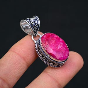 May include: A silver pendant with an oval-shaped, pink ruby gemstone set in a silver bezel. The pendant has an intricate, filigree design around the gemstone.