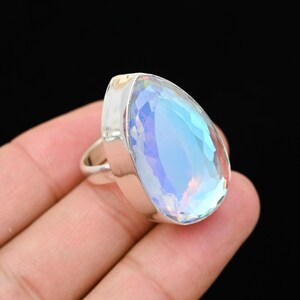 Puede incluir: Un anillo con una gran piedra preciosa en forma de pera, con una piedra iridiscente facetada. La piedra muestra una gama de colores, incluyendo azul, p&uacute;rpura y verde. El anillo tiene una banda plateada.