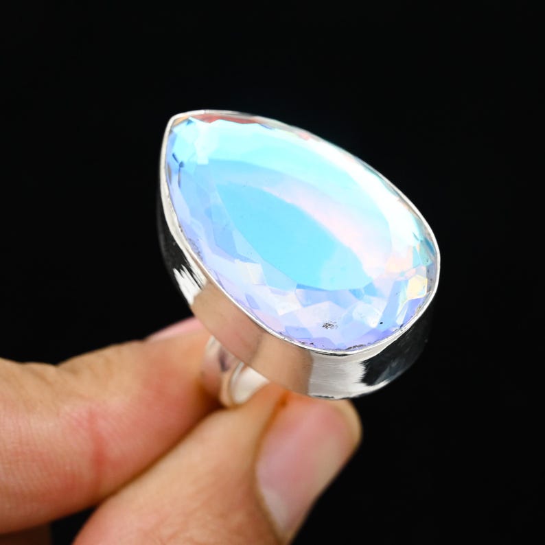Puede incluir: Anillo de plata con una gran gema en forma de pera. La piedra muestra una iridiscencia azul claro y est&aacute; facetada para reflejar la luz. El anillo se presenta sobre un fondo negro.