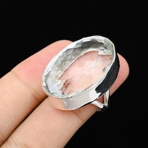 Op de afbeelding: Een ring met een heldere, ovaalvormige edelsteen met een gefacetteerd oppervlak. De ring heeft een zilveren band en zetting. De edelsteen wordt op zijn plaats gehouden door een bezel setting. Op een zwarte achtergrond.