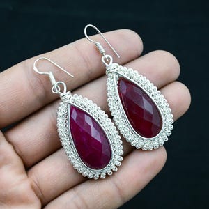 Op de afbeelding: Een paar zilveren oorbellen met een traanvormige, gefacetteerde, donkerroze edelsteen die is gezet in een zilveren draadkooi.