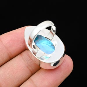 Puede incluir: Un anillo de plata con una gran gema ovalada de color azul claro. El anillo tiene una banda ancha y un engaste decorativo que sujeta la piedra. La superficie facetada de la gema captura la luz, creando un efecto brillante.