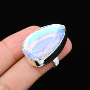 Puede incluir: Anillo de plata con una gran piedra preciosa iridiscente en forma de pera. La piedra refleja colores como el azul, el morado y el amarillo. El anillo est&aacute; sobre un fondo oscuro.
