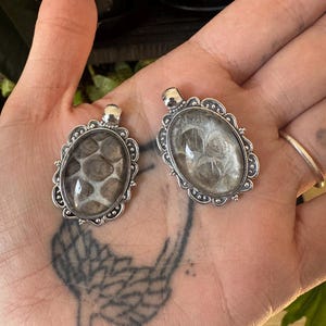 May include: Two oval pendants with silver-coloured filigree frames. One pendant features a snakeskin pattern, the other a wispy, cloud-like design. Both have a small loop at the top for a chain. The pendants are held in a hand.