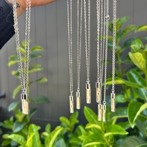 May include: A collection of silver-toned necklaces featuring clear, cylindrical glass vials as pendants. Each vial is capped with a metallic silver top and hangs from a delicate silver-coloured chain, displaying a small, light-coloured object inside.