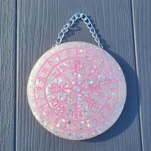 Peut inclure: Une suspension murale ronde, blanche et recouverte de paillettes avec un motif circulaire rose et des runes autour du bord. La suspension est attachée à une chaîne argentée.