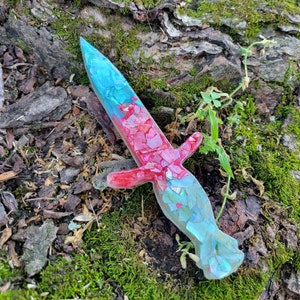 Może przedstawiać: Czerwono-niebieski, przypominający sztylet, przycisk do papieru z żywicy o błyszczącym wykończeniu. Przycisk do papieru wykonany jest z małych kawałków szkła i leży na mchu.