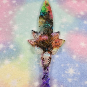 May include: A colorful resin dagger with a flower-shaped handle and a glitter finish. The blade is a gradient of green, yellow, and purple.