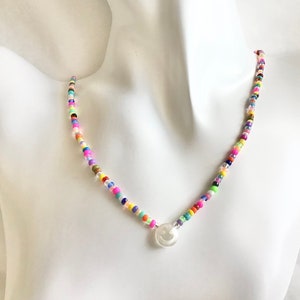 Könnte beinhalten: Eine bunte Perlenkette mit einem weißen Smiley-Anhänger. Die Perlen sind klein und rund und in einem zufälligen Muster angeordnet. Die Kette besteht aus verschiedenen Farben, darunter Pink, Blau, Grün, Gelb und Orange.