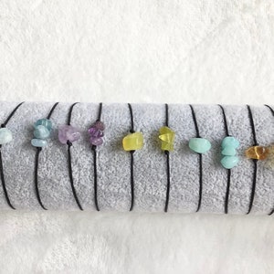 Könnte beinhalten: Neun Armbänder mit verschiedenen farbigen Edelsteinen auf schwarzer Schnur. Die Edelsteine sind Aquamarin, Amethyst, Citrin und Bernstein.