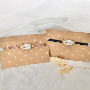 Könnte beinhalten: Zwei handgefertigte Armbänder mit einem Muschelanhänger. Die Armbänder befinden sich auf einer braunen Karte mit weißen Sternen und dem Text "HANDMADE".