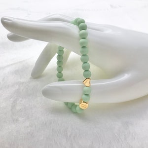 Puede incluir: Un brazalete de cuentas de color verde claro con abalorios de coraz&oacute;n y letras doradas. El brazalete est&aacute; en una mano de maniqu&iacute; blanca.