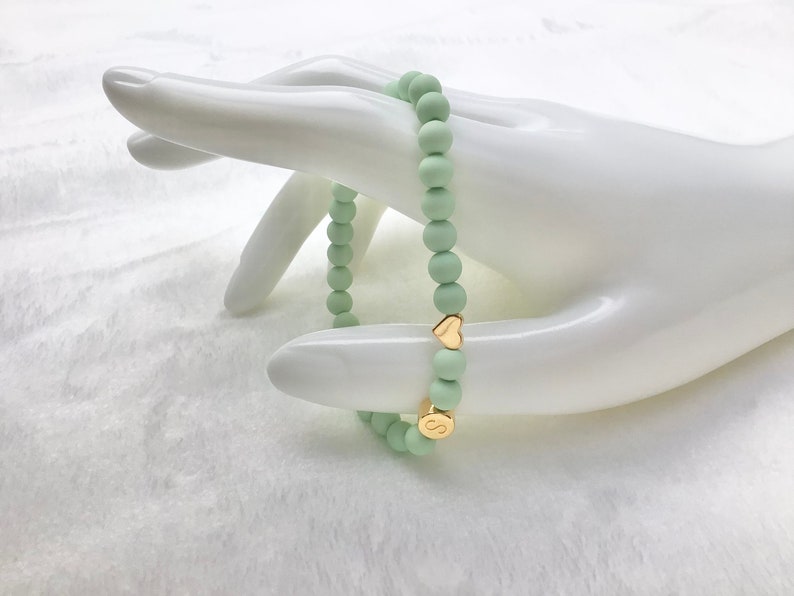 Puede incluir: Un brazalete de cuentas de color verde claro con abalorios de coraz&oacute;n y letras doradas. El brazalete est&aacute; en una mano de maniqu&iacute; blanca.