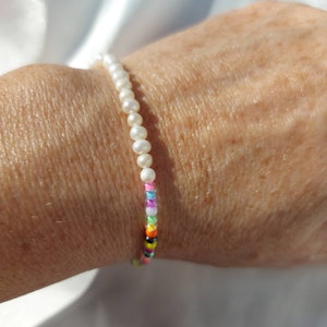 Könnte beinhalten: Ein weißes Perlenarmband mit einem bunten Perlenakzent. Die Perlen sind klein und schimmernd und erzeugen einen Regenbogeneffekt.