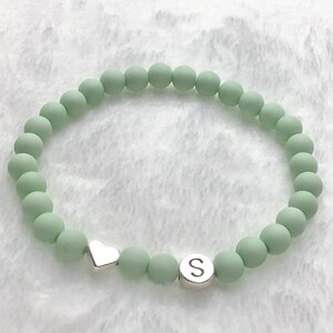 Puede incluir: Un brazalete de cuentas verde claro con un dije de coraz&oacute;n plateado y un dije plateado con la letra "S".
