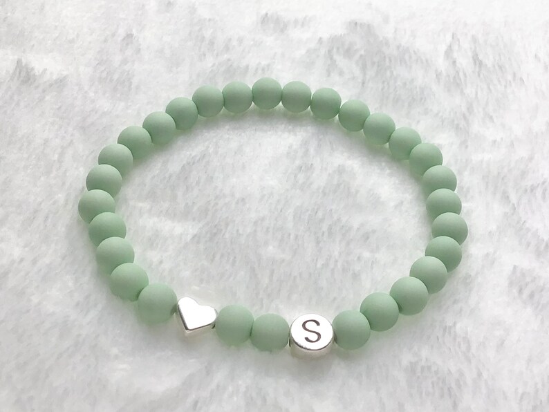 Puede incluir: Un brazalete de cuentas verde claro con un dije de coraz&oacute;n plateado y un dije plateado con la letra "S".