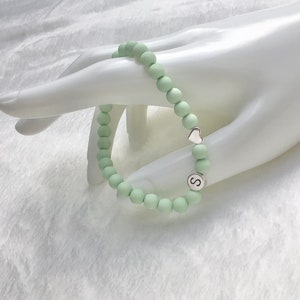 Puede incluir: Un brazalete de cuentas de color verde claro con un dije de coraz&oacute;n plateado y un dije plateado con la letra 'S'.