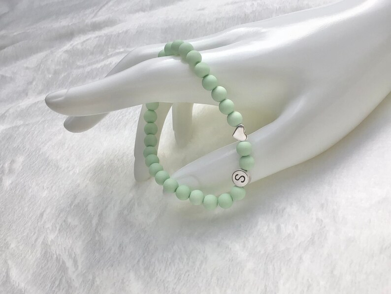 Puede incluir: Un brazalete de cuentas de color verde claro con un dije de coraz&oacute;n plateado y un dije plateado con la letra 'S'.