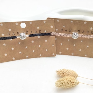 Könnte beinhalten: Zwei handgefertigte Freundschaftsarmbänder mit Eulenanhängern. Die Armbänder befinden sich auf braunem Karton mit weißen Sternen und dem Text "HANDMADE".