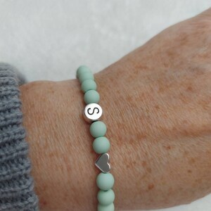Puede incluir: Un brazalete de cuentas de color verde claro con un dije de plata que tiene la letra "S" y un dije de coraz&oacute;n de plata.