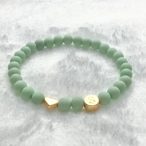 Puede incluir: Un brazalete de cuentas verdes con un dije de coraz&oacute;n dorado y una cuenta dorada con la letra "S" en ella.