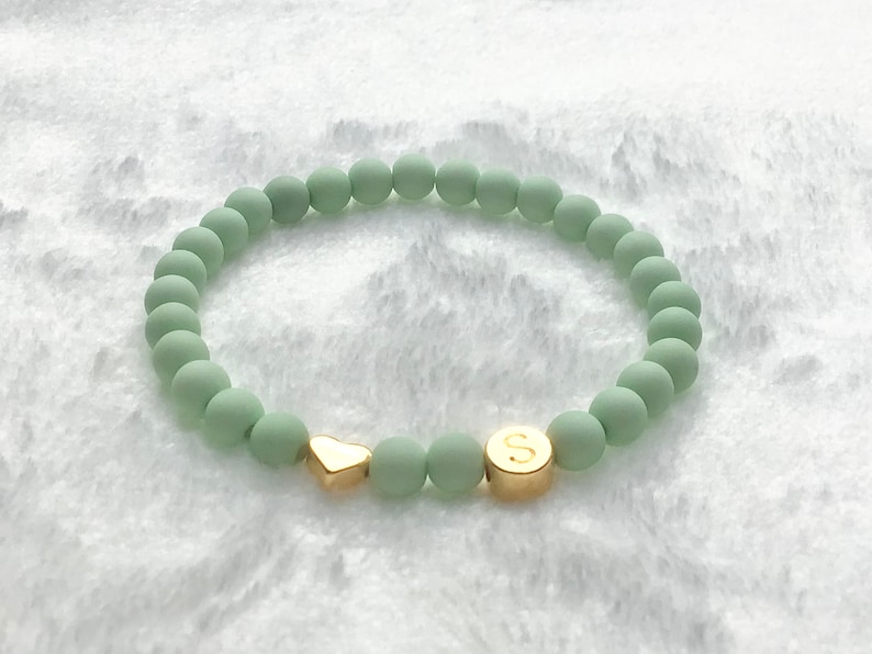 Puede incluir: Un brazalete de cuentas verdes con un dije de coraz&oacute;n dorado y una cuenta dorada con la letra "S" en ella.