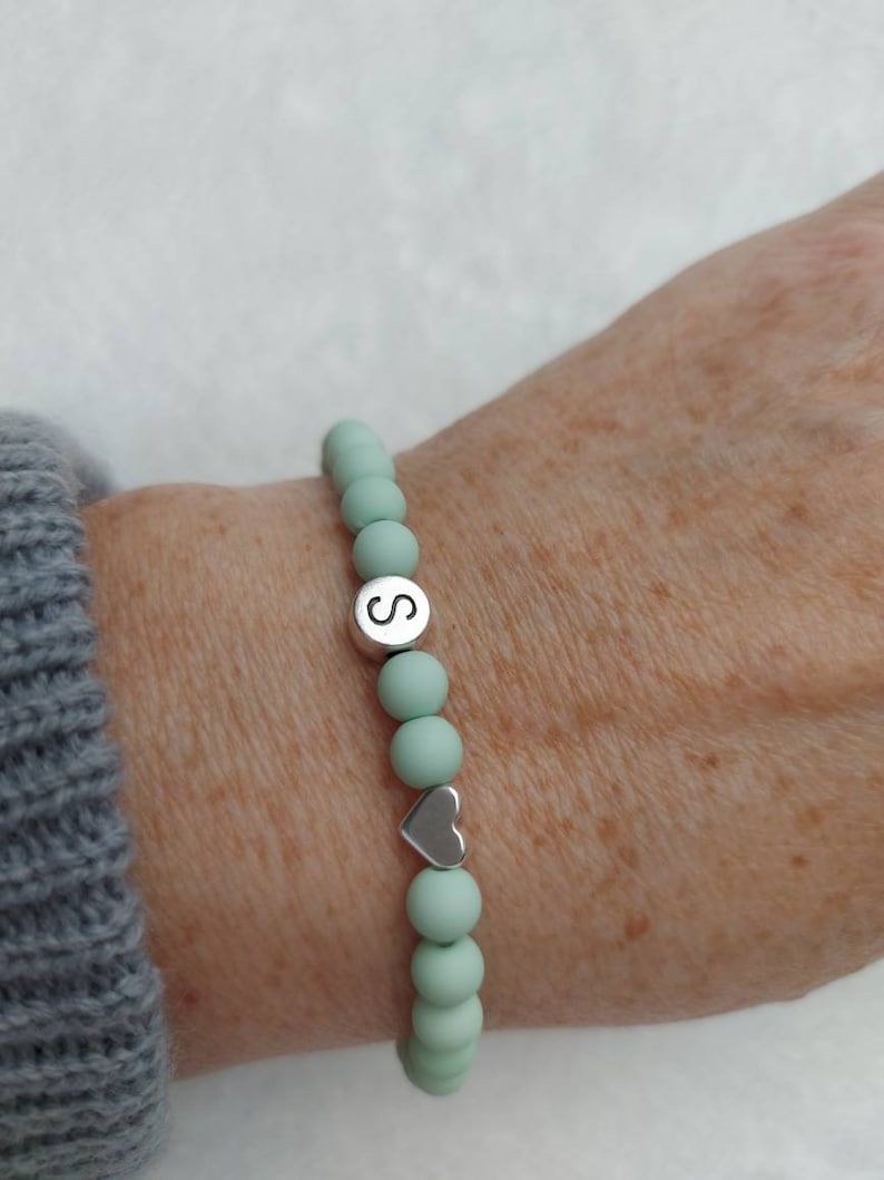 Puede incluir: Un brazalete de cuentas de color verde claro con un dije de plata que dice "S" y un dije de coraz&oacute;n de plata.