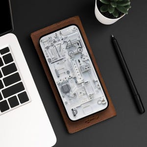 Op de afbeelding: Een bruine leren telefoonhoes met een witte telefoon erin. Het telefoonscherm toont een klok met de tijd 17:21 en de datum zo 28. De achtergrond van de telefoon is een wit en grijs abstract ontwerp.