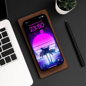 Könnte beinhalten: Eine braune Leder-Handyhülle mit einem rosa und blauen Sonnenuntergang und Palmen auf dem Bildschirm. Der Bildschirm des Telefons zeigt die Zeit 23:50 und das Datum Fr 8. Der Text "BTC $44,124.01" ist ebenfalls auf dem Bildschirm zu sehen.