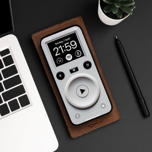May include: A brown leather case with a white phone inside. The phone screen displays a digital clock showing 21:59 and the date Sunday, 7 April. The phone has a large round button with a play icon in the center.