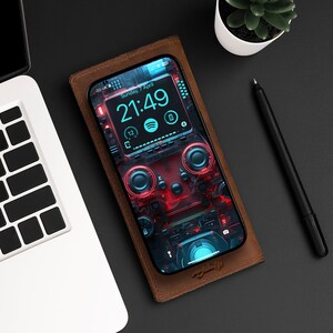May include: A smartphone with a gaming-themed wallpaper rests on a brown leather phone case. The phone displays the time, date, and app icons. A laptop, pen, and small plant are also visible on a dark surface.