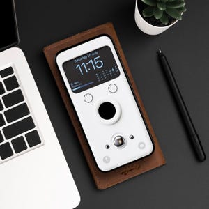 Puede incluir: Una funda de teléfono blanca con una cubierta de cuero marrón. La pantalla del teléfono muestra la fecha y la hora, "Sábado 20 de julio 11:15".