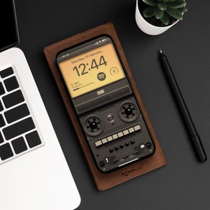 Puede incluir: Una funda de teléfono de cuero marrón con un diseño de cinta de casete de estilo vintage. La pantalla del teléfono muestra un reloj digital que muestra la hora 12:44 y la fecha sábado 10 de febrero. El teléfono está cargado al 29%.