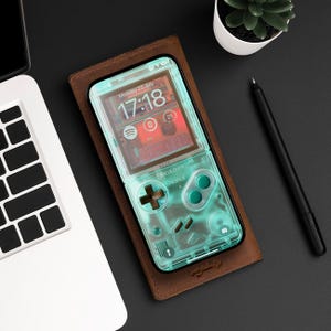 Pode incluir: Uma capa de telefone de couro marrom com uma capa de telefone transparente azul claro dentro. A capa do telefone tem um design de controlador de jogo com botões e uma tela que mostra a hora e a data. O texto na tela diz "Segunda-feira, 22 de julho 17:18".