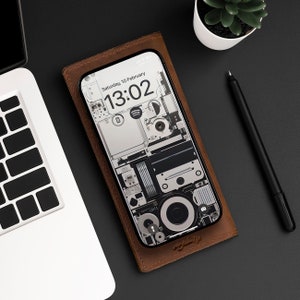 Op de afbeelding: Een bruine leren telefoonhoes met een witte en grijze telefoon erin. Het telefoonscherm toont een klok met de tijd 13:02 en de datum zaterdag 10 februari. De telefoonhoes heeft een geometrisch ontwerp.