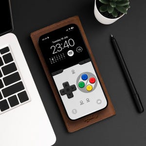 May include: A brown leather phone case with a white phone inside. The phone screen displays a digital clock with the time 23:40 and the date Tuesday 16 July. The phone screen also shows a video game controller icon with four colored buttons.