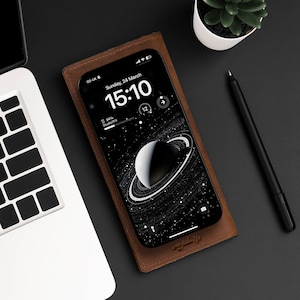 Pode incluir: Uma capa de telefone de couro marrom com um telefone preto dentro. A tela do telefone exibe um fundo espacial preto e branco com um planeta e anéis. O horário é 15:10 e a data é domingo, 24 de março.
