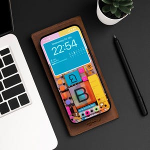 Pode incluir: Uma capa de telefone de couro marrom com um design abstrato colorido e uma tela de telefone branca que mostra a hora "22:54" e a data "quarta-feira, 24 de julho".