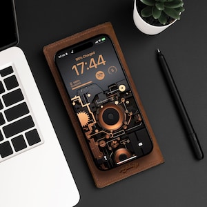Puede incluir: Una funda de teléfono de cuero marrón con un teléfono negro en su interior. La pantalla del teléfono muestra un reloj con la hora 17:44 y el texto "95% cargado". El fondo del teléfono es una ilustración estilizada de engranajes y circuitos.