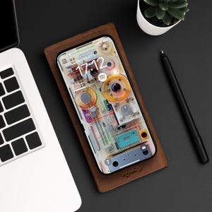Peut inclure: Un étui de téléphone en cuir marron avec un téléphone transparent à l'intérieur. L'écran du téléphone affiche une horloge numérique indiquant l'heure 17:12 et la date du lundi 22 juillet. L'arrière-plan du téléphone est un motif abstrait coloré.