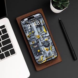 以下が含まれることがあります： 茶色の革製の携帯電話ケースの中に黒い携帯電話が入っています。携帯電話の画面には、21:48 と表示されたデジタル時計と、7月25日木曜日という日付が表示されています。携帯電話の背景は、黄色と灰色のディテールが施された回路基板のグラフィックデザインです。