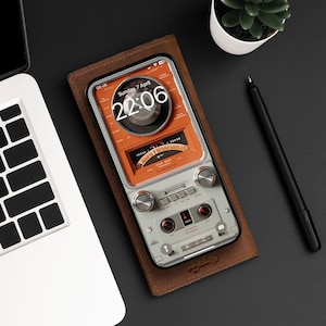 Può includere: Una custodia per telefono in pelle marrone con un design radio vintage. Lo schermo del telefono mostra un orologio digitale con l'ora 22:06 e la data domenica 7 aprile. La custodia ha un bordo e pulsanti argentati.