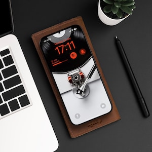May include: A brown leather phone case with a smartphone inside. The phone screen displays a black and white image of a record player with a red needle. The time on the phone screen is 17:11.